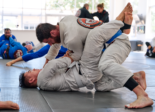Two adult students practicing Brazilian Jiu Jitsu grappling techniques on padded mats at Legacy Brazilian Jiu Jitsu. One student in a white gi works from a top position while the other defends from the ground using guard control. Additional students are visible training in the background inside a bright, open martial arts academy