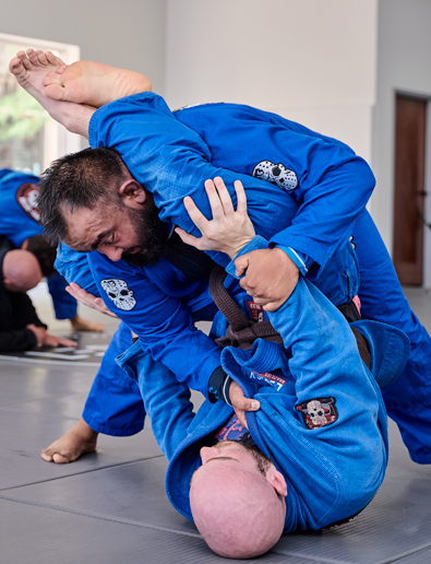 Two adult students practicing Brazilian Jiu Jitsu sparring techniques on padded mats at Legacy Brazilian Jiu Jitsu. Both are wearing blue gis as one student works from a standing position while the other executes a guard technique from the ground. Other students can be seen training in the background inside a bright martial arts studio.