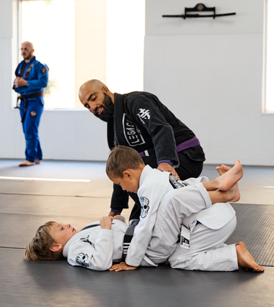 Two children practicing Brazilian Jiu Jitsu during a class at Legacy Brazilian Jiu Jitsu. One child in a white gi is positioned on the mat while another practices from the top position under the guidance of an instructor in a black gi with a purple belt. Another instructor wearing a blue gi stands in the background inside a bright martial arts training studio.