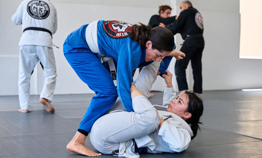 Two women practicing Brazilian Jiu Jitsu techniques during a class at Legacy Brazilian Jiu Jitsu. A student in a blue gi works from a standing position while a student in a white gi practices guard control from the mat. Other students train in the background inside a bright martial arts academy.